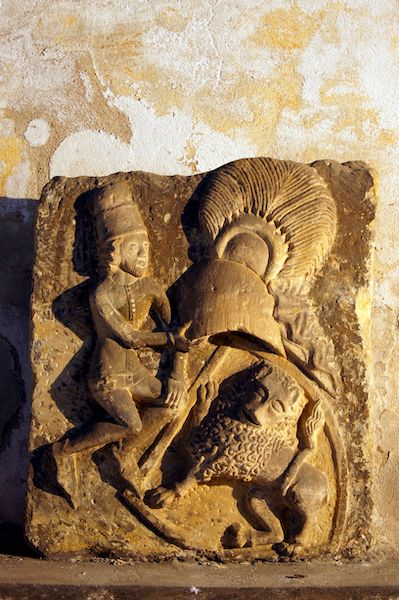 Eine in Stein gehauene menschliche Figur mit einem Helm mit Federschmuck in der Hand, daneben ein Löwe