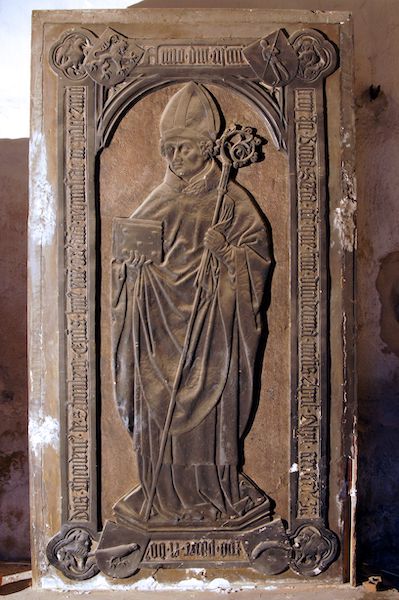 Sandsteinplatte mit einer Bischofsfigur rechteckig umrahmt von Schrift und Wappen an den Ecken